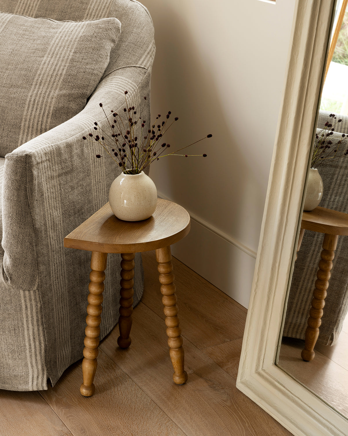 The Makerspalm Alice Honey Wood Stool, a compact ash wood accent with three spindle legs, displays a round beige vase with dried branches beside a gray-striped armchair and large framed mirror on a wooden floor, Vendixs