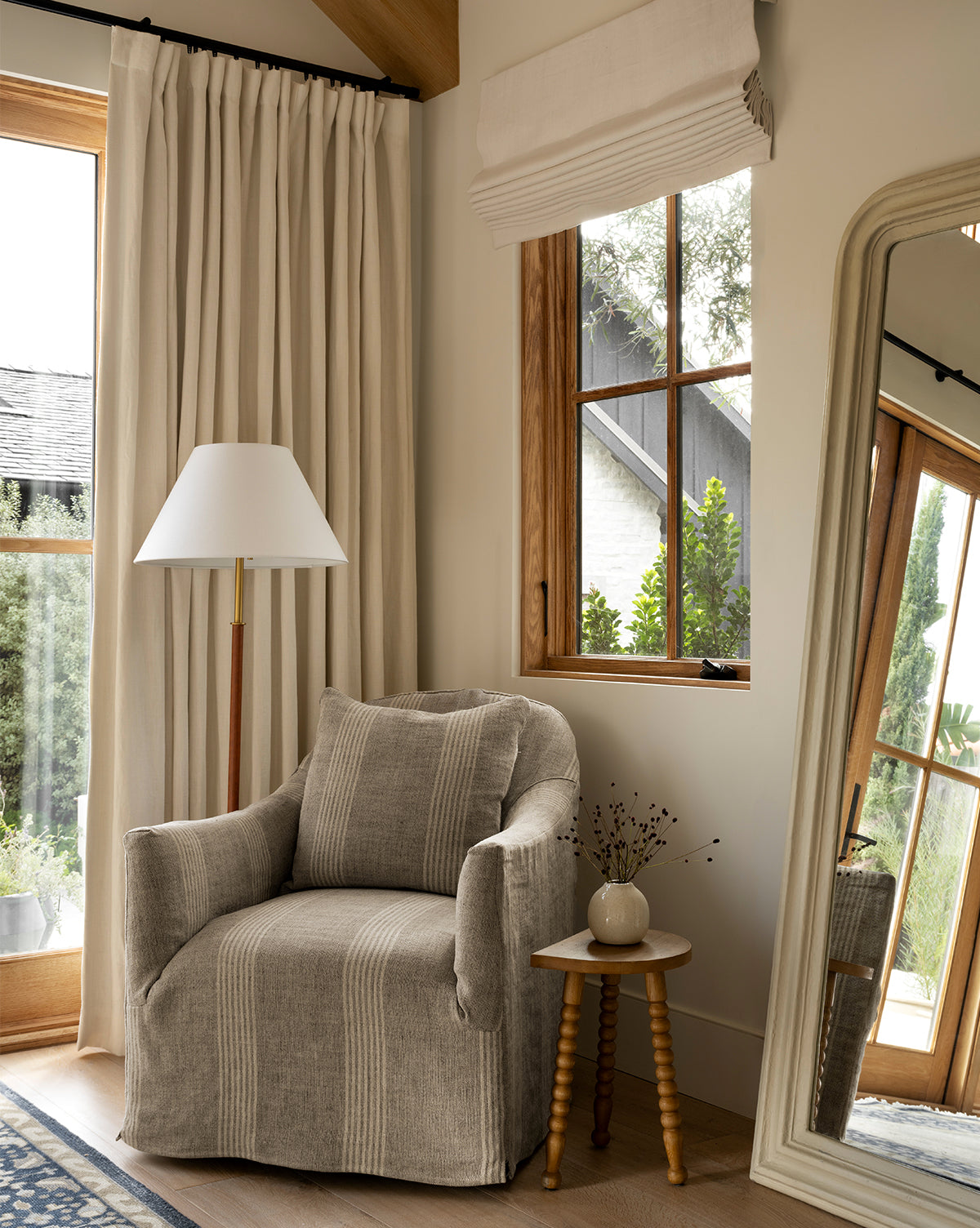 A cozy corner features the Trudeaux Slipcover Swivel Chair by Rowe Fine Furniture, Inc., a striped pillow, wooden side table with vase, tall floor lamp, large mirror, cream curtains, and a window of greenery bathed in warm natural light—all styled with touches from Vendixs