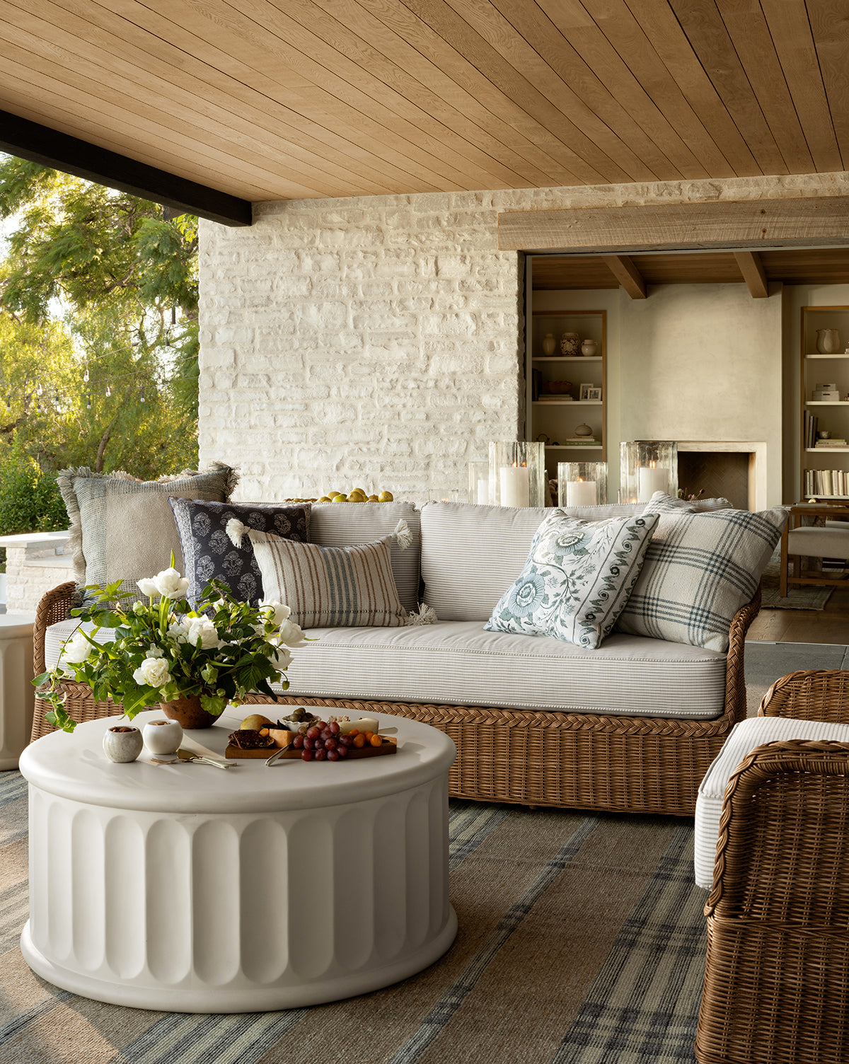 A cozy outdoor patio features the Haviland Outdoor Sofa with Perennials Taupe Stripe 88, patterned cushions, a white round coffee table with flowers and snacks, a rug over wooden flooring, and greenery visible outside by Vendixs