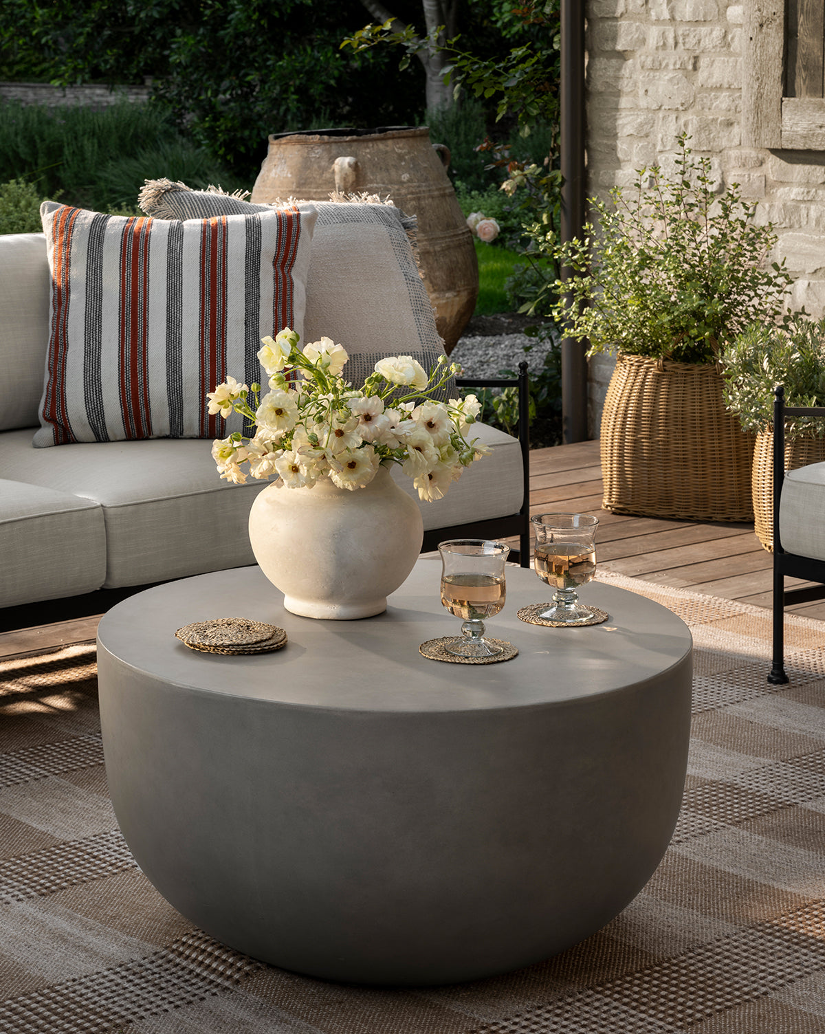 A modern patio showcases a round gray coffee table with a Nola Paper Mache Vase holding white flowers and two glasses, while a beige sofa with striped pillows and lush potted plants create a cozy, inviting outdoor space, by Vendixs