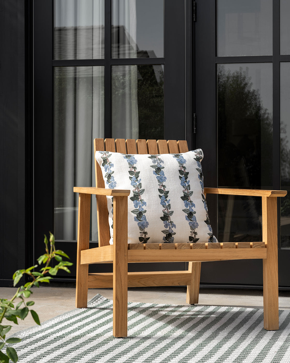 The Gilda Outdoor Lounge Chair, crafted from teak wood with a white cushion featuring blue floral patterns, sits on a striped rug before black-framed glass doors. A leafy plant appears in the foreground, by Vendixs
