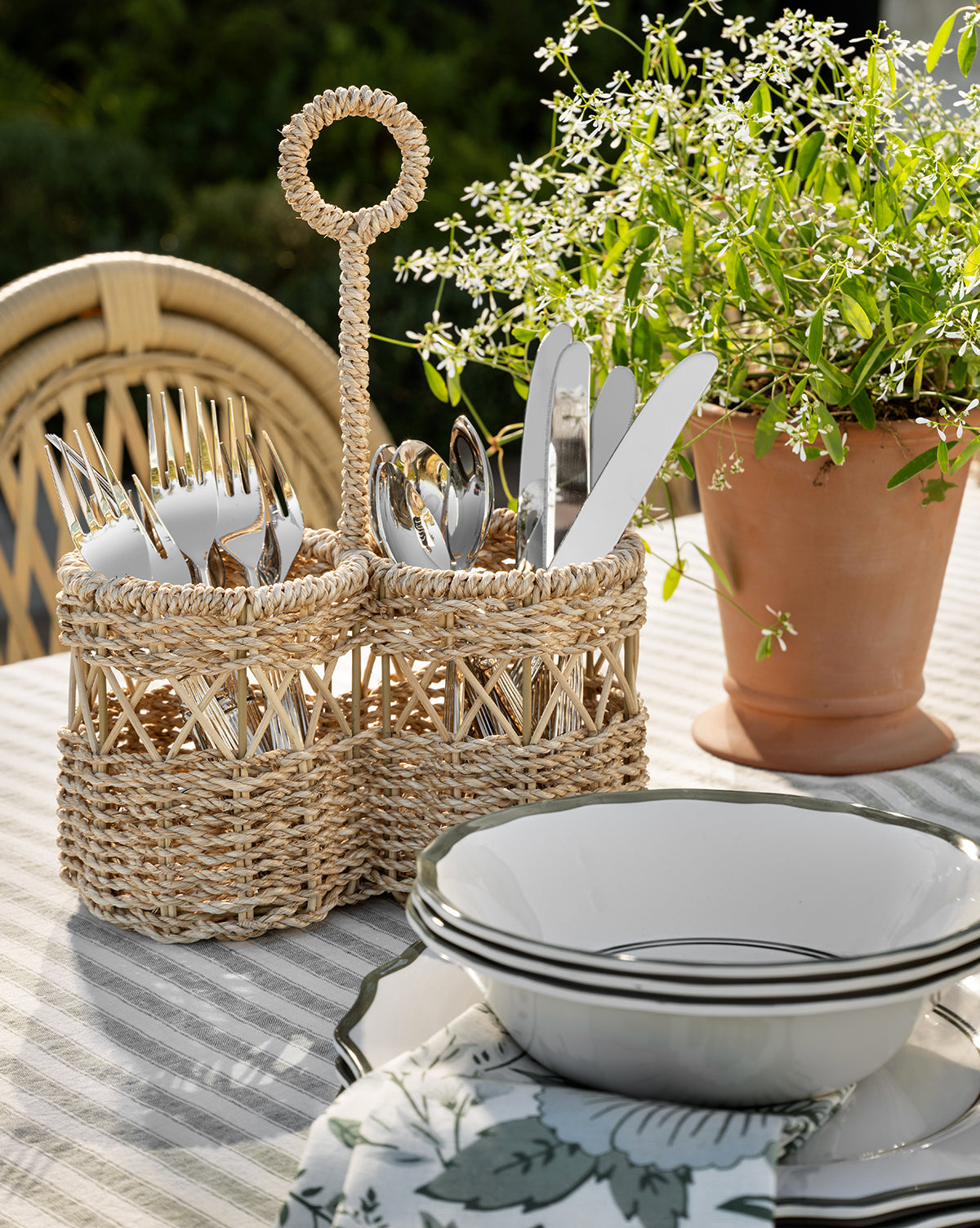 The Stokes Woven Caddy with silverware sits on a striped tablecloth beside stacked white plates and a potted plant, creating an inviting outdoor dining setting, by Vendixs