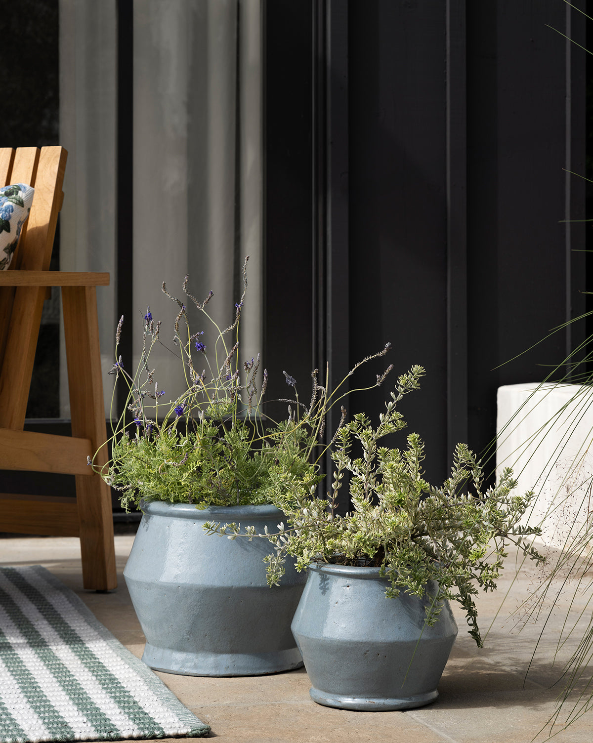 Two UNITED POTTERIES SAIGON Corsica Pots with green and lavender plants sit on a patio beside a wooden chair and striped rug, with glass doors framed in black metal in the background. Vendixs
