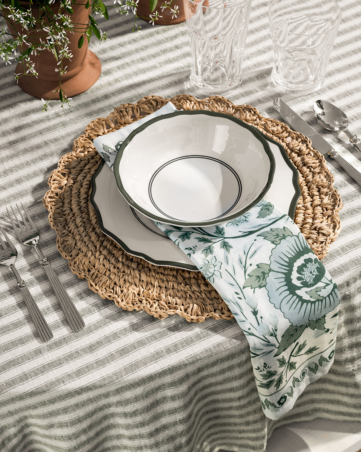 A table setting with a woven placemat, scalloped edge white plate, Alcott Melamine Bowls (Set of 4) with green rims, floral napkin, striped tablecloth, silverware, clear glasses, and potted plants in the background by Vendixs
