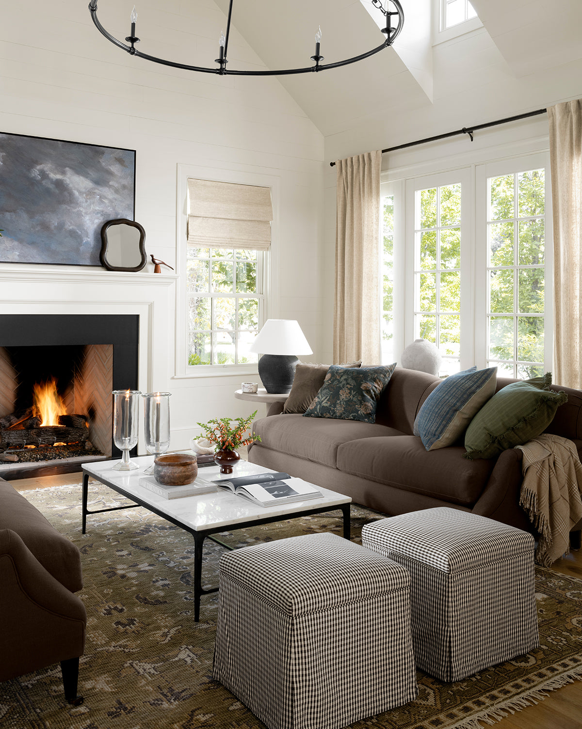 A cozy living room with a glowing fireplace, brown sofas, plaid ottomans, and a Wayland Hand-Knotted Wool Rug. Large windows let in sunlight, while neutral curtains frame views of trees outside, by Vendixs