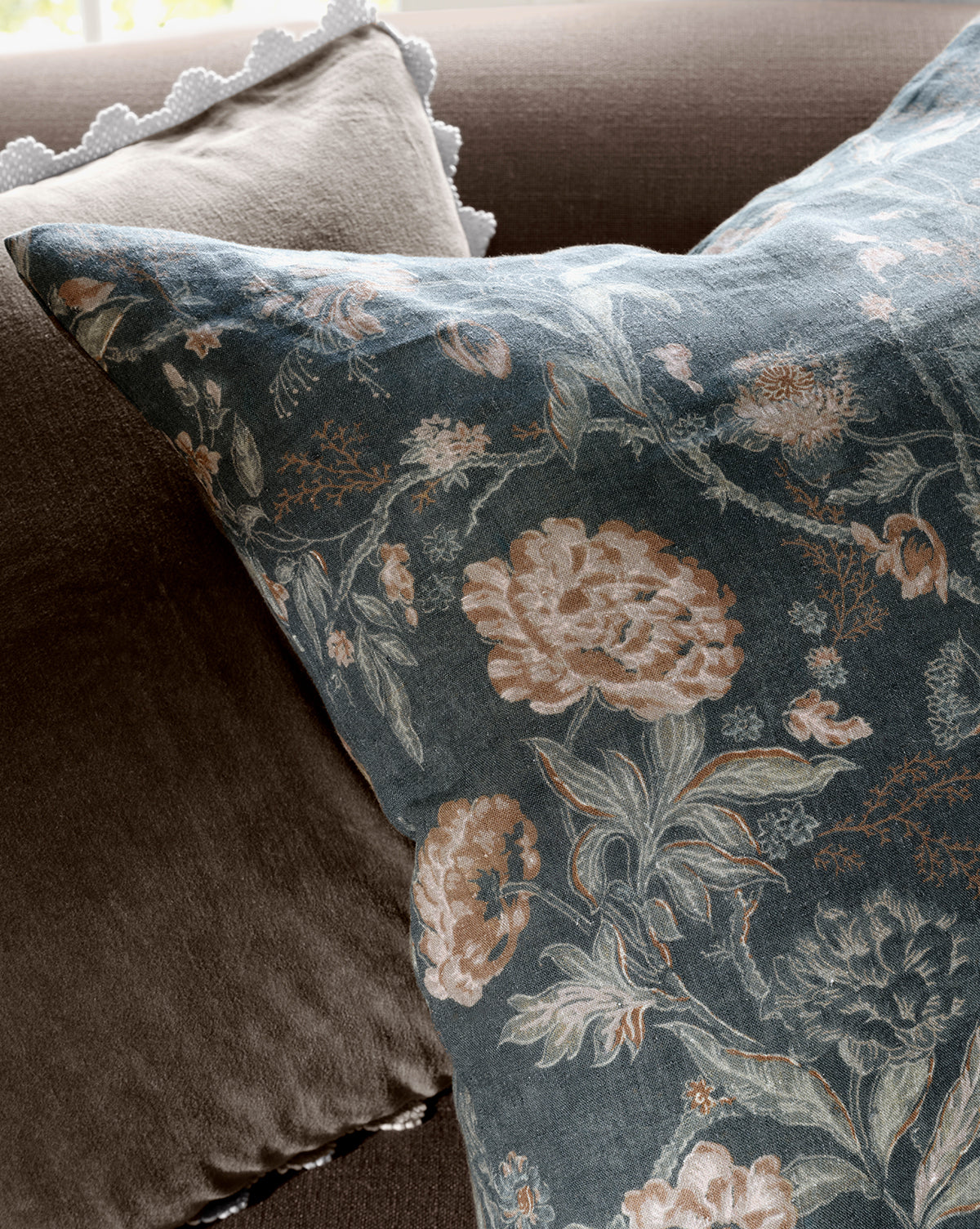 A close-up of the Hestercombe Pillow Cover, showcasing vintage-inspired muted pink and beige flowers on a dark background, paired with a plain brown pillow and a white scalloped-edged cushion in the background, by Vendixs