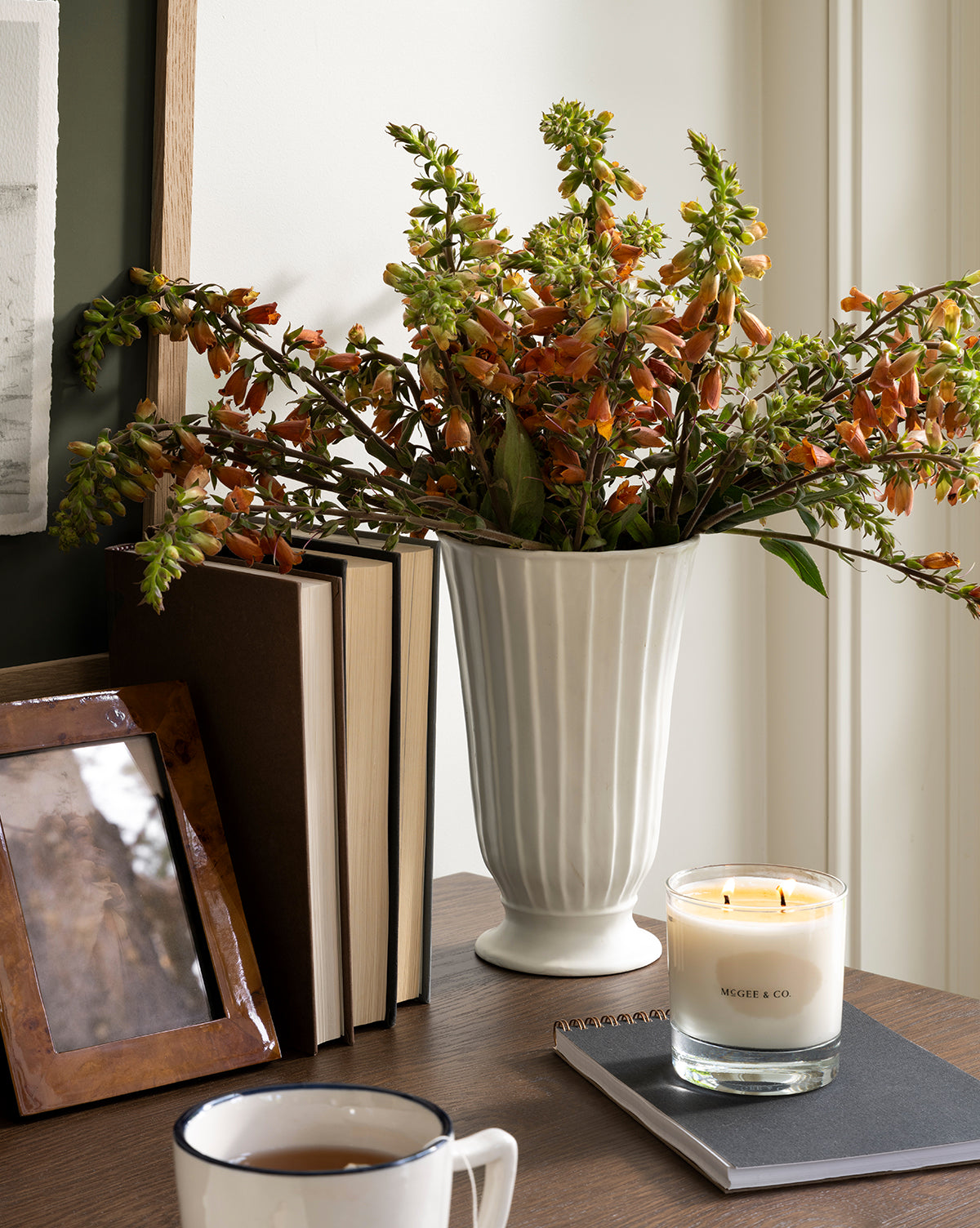 The Delphi Footed Vase, crafted from white stoneware and filled with orange flowers, rests on a wooden table beside a candle, closed notebook, mug, framed photo, and upright books by Vendixs
