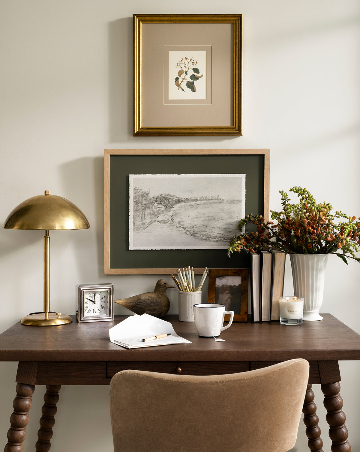 A tidy wooden desk with a beige chair, gold lamp, books, mug, and white vase with flowers adds charm to this decor. Lit candles glow while two Cove in Charcoal sketches in light oak frames hang above the desk, by Vendixs