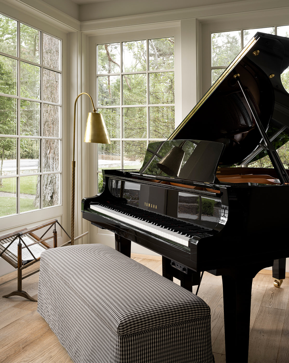 A black Yamaha grand piano sits in a bright room with large windows, a Latimer Bench in front, a brass floor lamp beside it, and trees visible outside, by Vendixs