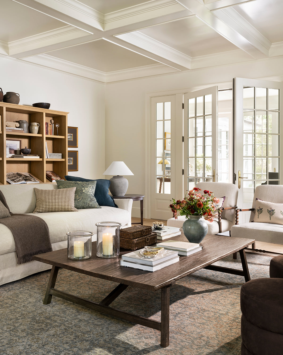 A cozy living room features a beige sofa, cushioned chairs, and the Lockwood Coffee Table. Shelves display decor, French doors fill the space with natural light, and the table holds candles, books, and a vase of flowers by Vendixs