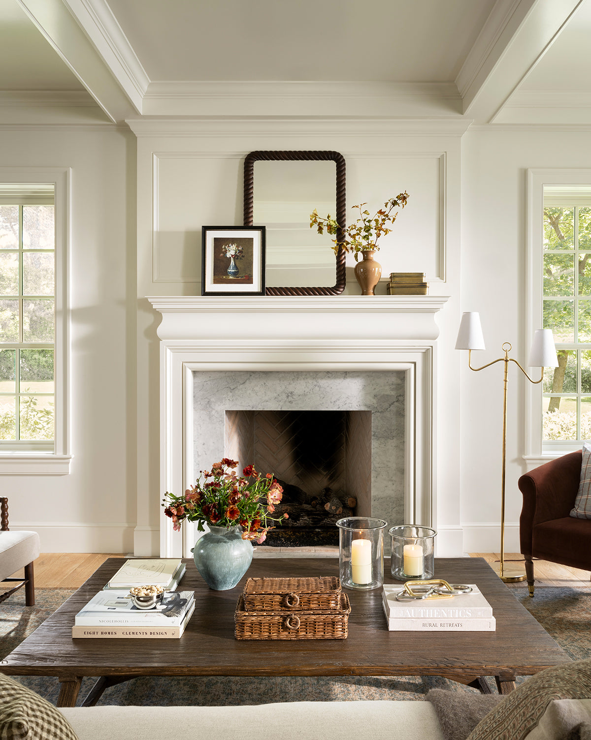 Elegant living room with a white fireplace, Mayweather Wall Mirror with a rope-inspired frame above the mantel, framed art, and a vase with branches. A dark red walnut coffee table displays books, candles, flowers, and a wicker tray. Sunlight streams through large windows, by Vendixs
