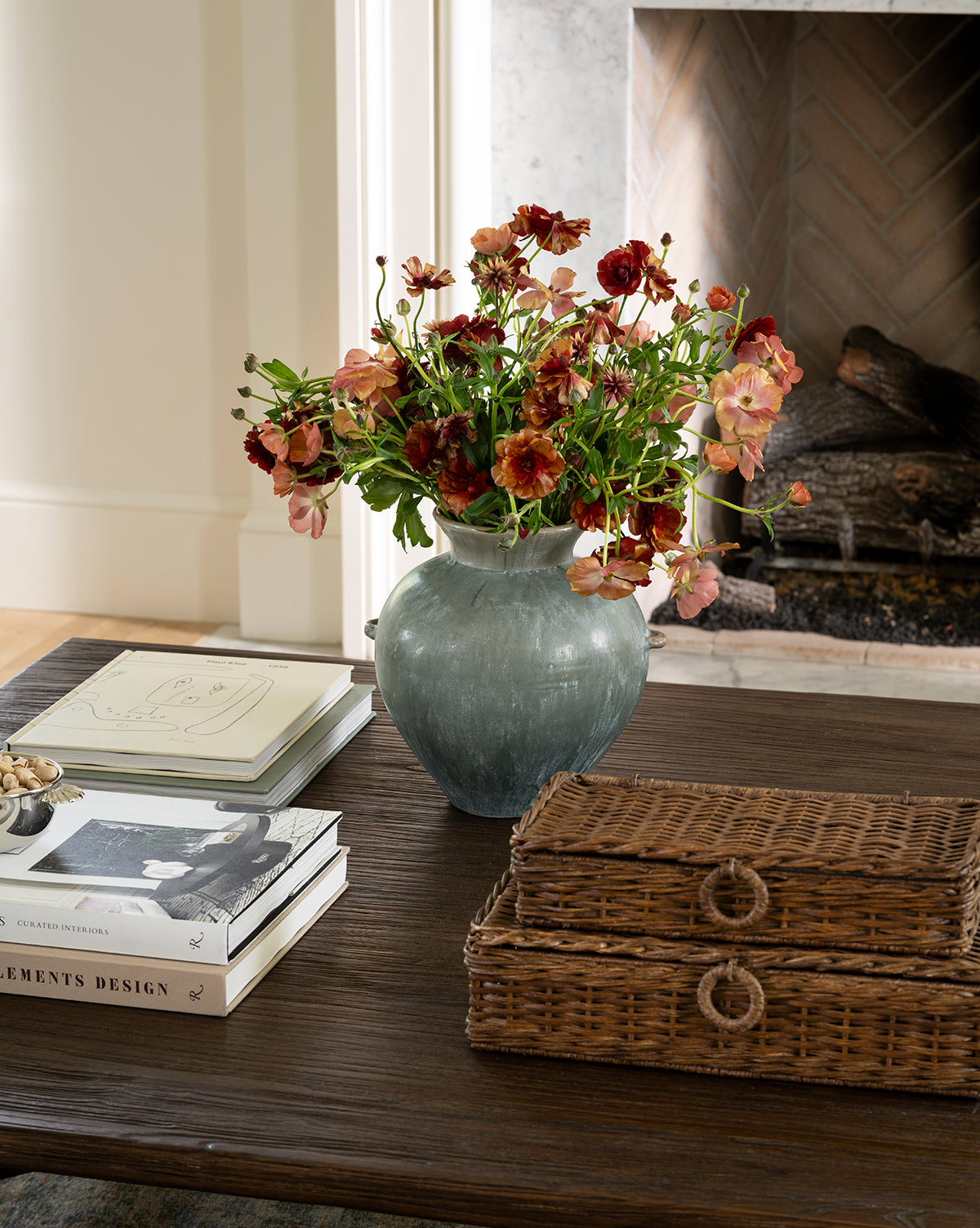 A Beatrix Vase brimming with red and pink blooms sits on a dark wood coffee table next to wicker boxes, books, and papers, with a fireplace behind it—showcasing the vintage-inspired handcrafted artistry of the Beatrix Vase, by Vendixs