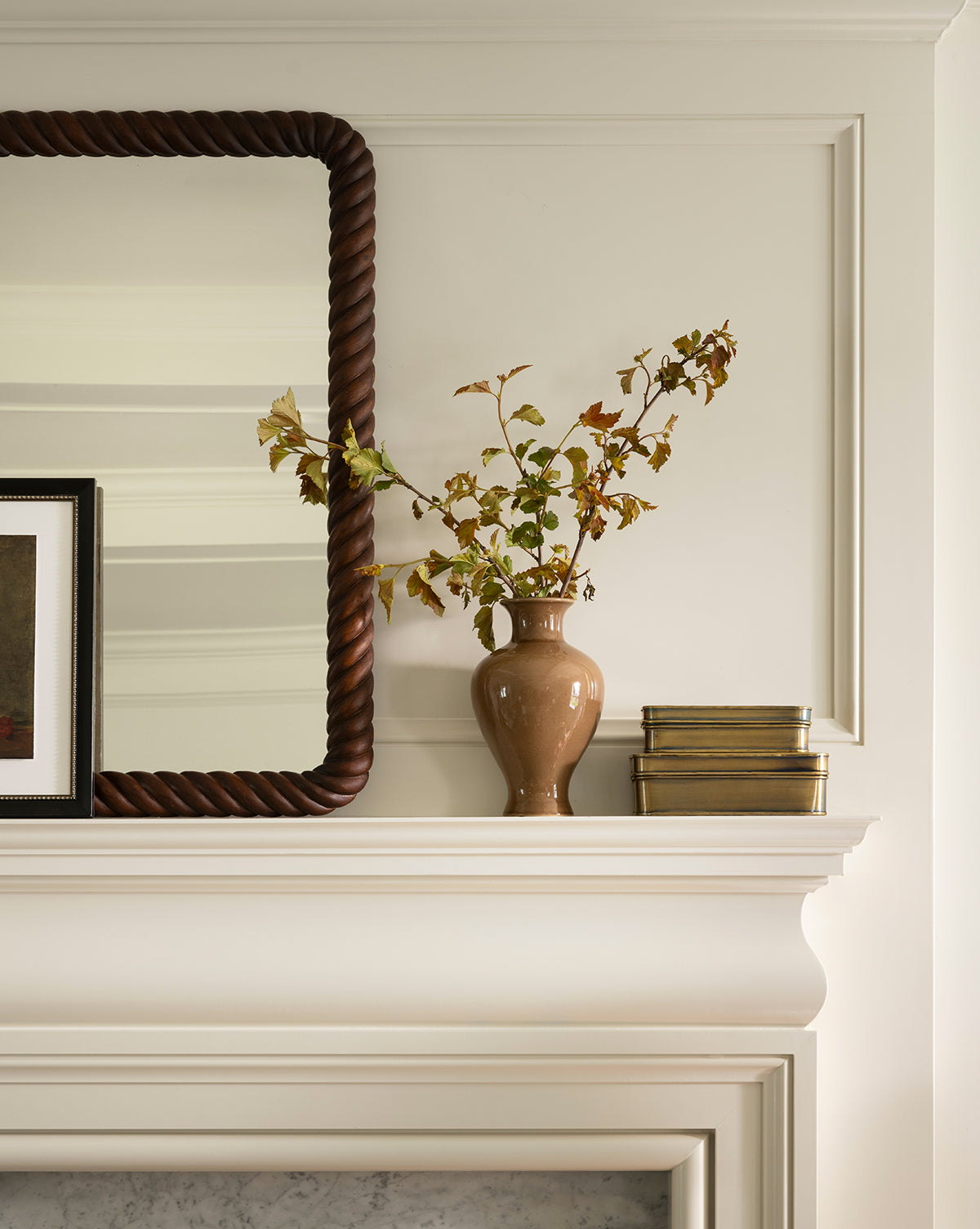 The Natalia Vase, filled with leafy branches, sits on a white mantel next to two gold boxes, with a large mirror in a twisted dark frame and a framed picture beside it, by Vendixs