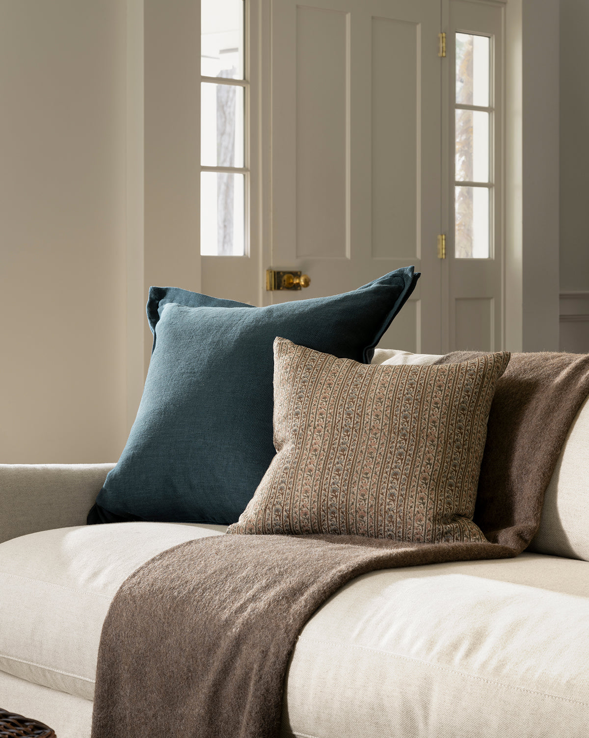 A cozy beige sofa with a dark blue pillow, the Wakefield Pillow Cover in a patterned brown design, and a brown throw blanket, set in a softly lit room with white paneled doors and large windows in the background, by Vendixs
