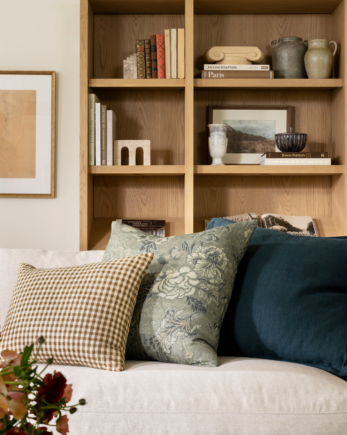 A beige sofa with patterned and solid throw pillows, including the Liam Double Flange Pillow Cover, sits in front of a wooden bookshelf filled with books and decor. Framed artwork hangs nearby, by Vendixs