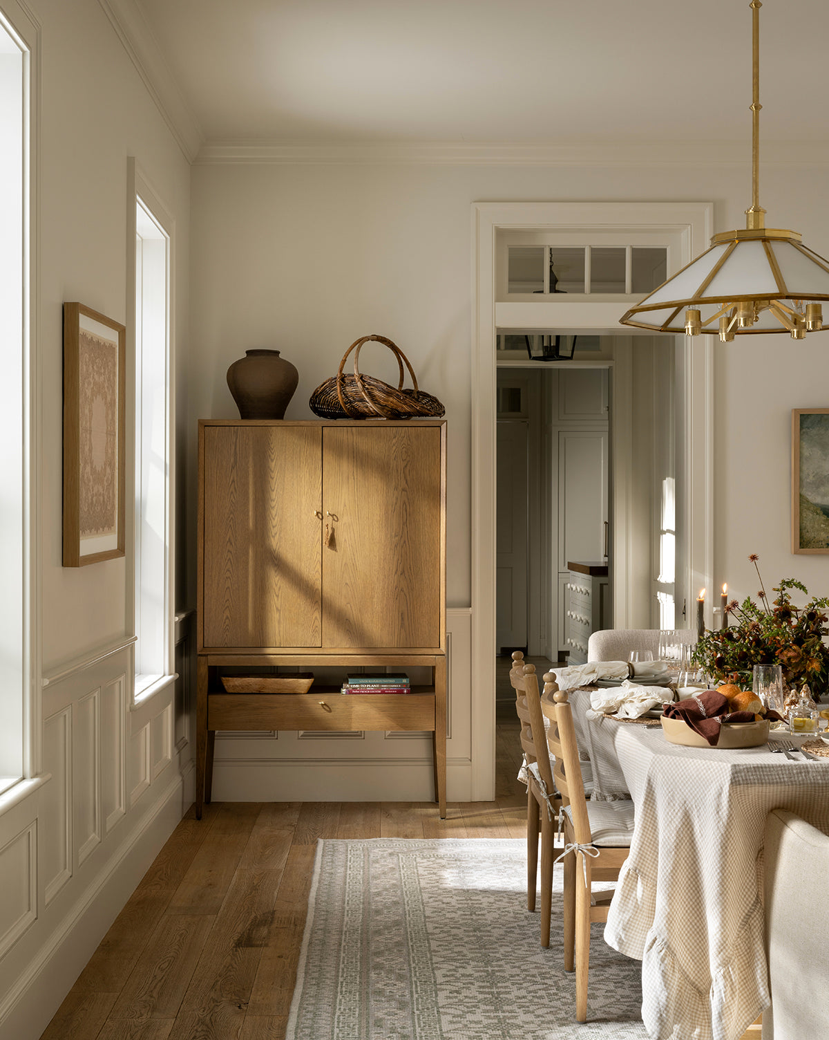 A bright dining room features a wooden table set for a meal with chairs, while the Orsino Hand-Knotted Rug adds warmth beneath. A light wood cabinet and large windows complete the cozy, neutral space by Vendixs