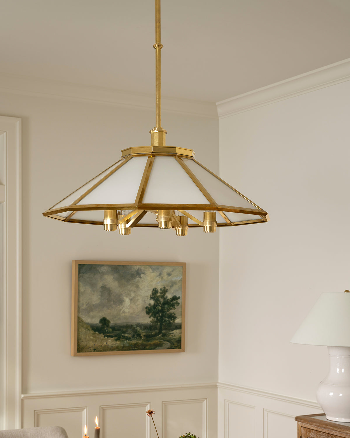 The Knightley Chandelier with its geometric antique brass design hangs above a neutral-toned room, while a framed landscape artwork and a white table lamp add contrast to the light-colored walls, by Vendixs