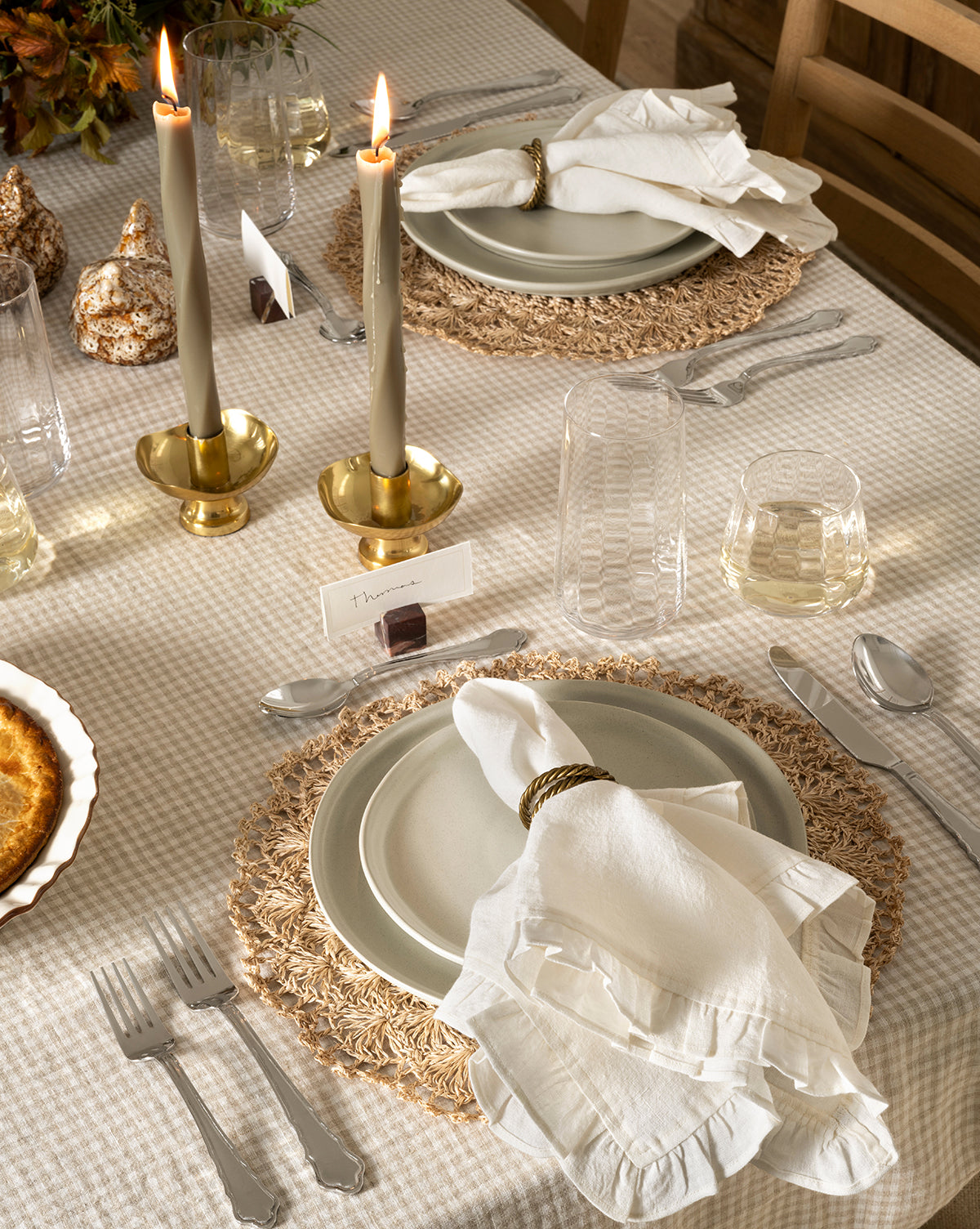 A set dining table with neutral-toned plates, white cloth napkins and rings, woven placemats, clear glasses, the Artois Flatware (Set of 5), brass candlesticks with lit candles, place cards, and a checked tablecloth by Vendixs