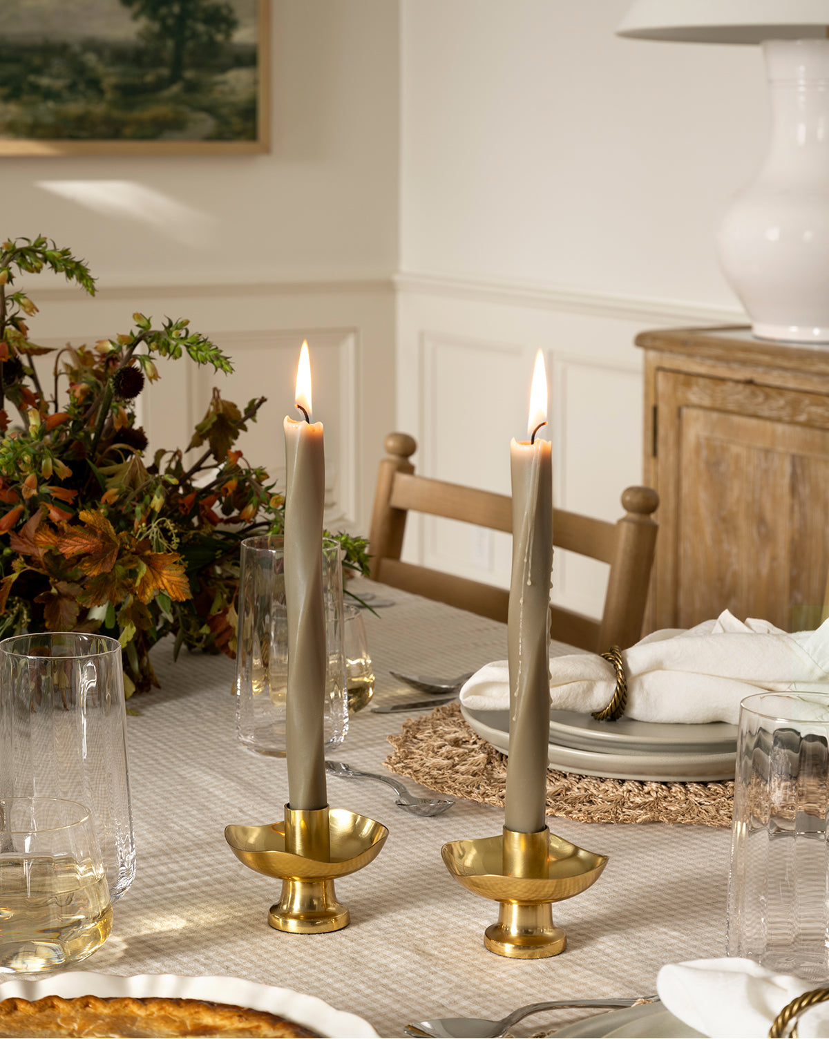 Two lit Errol Wax Tapers (Set of 6) in brass holders sit on a dining table with glassware, woven placemat, white napkins, and a fall floral arrangement. A sideboard and framed art are visible in the background, by Vendixs