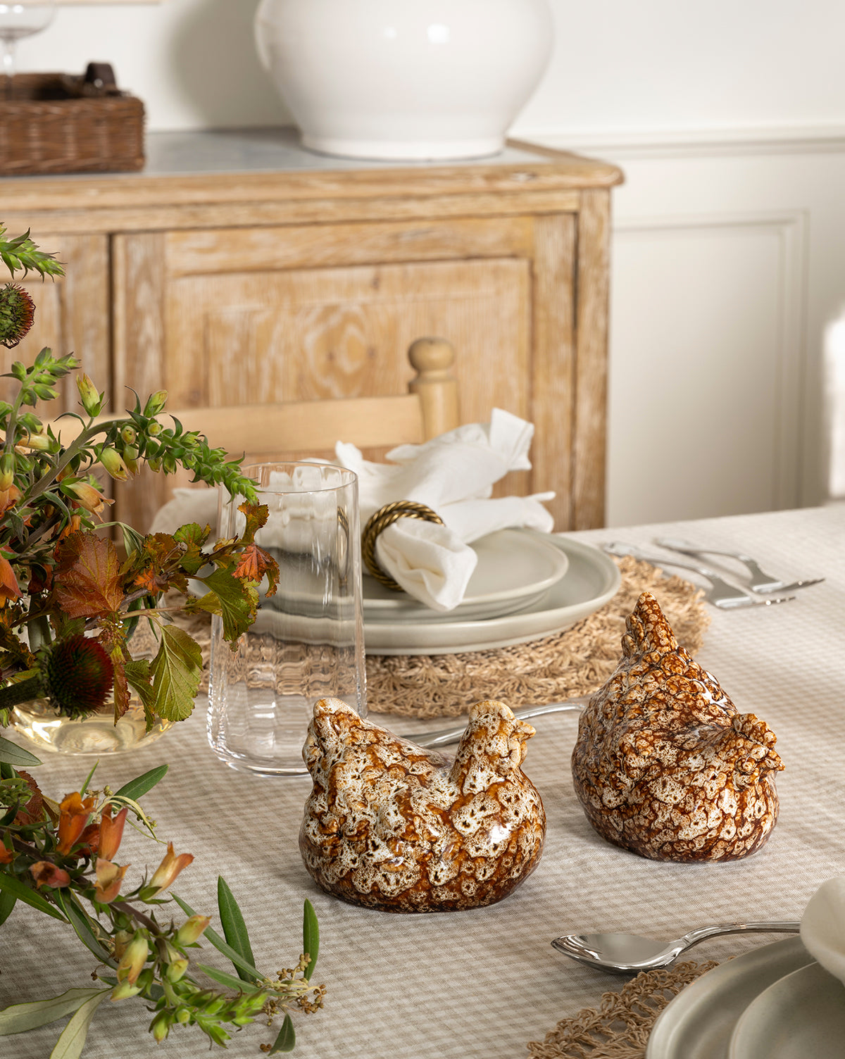 A dining table set with neutral plates, glasses, and napkins displays artisanal charm with a Stoneware Hens (Set of 2) centerpiece amid autumn foliage; a wooden sideboard appears in the background, by Vendixs