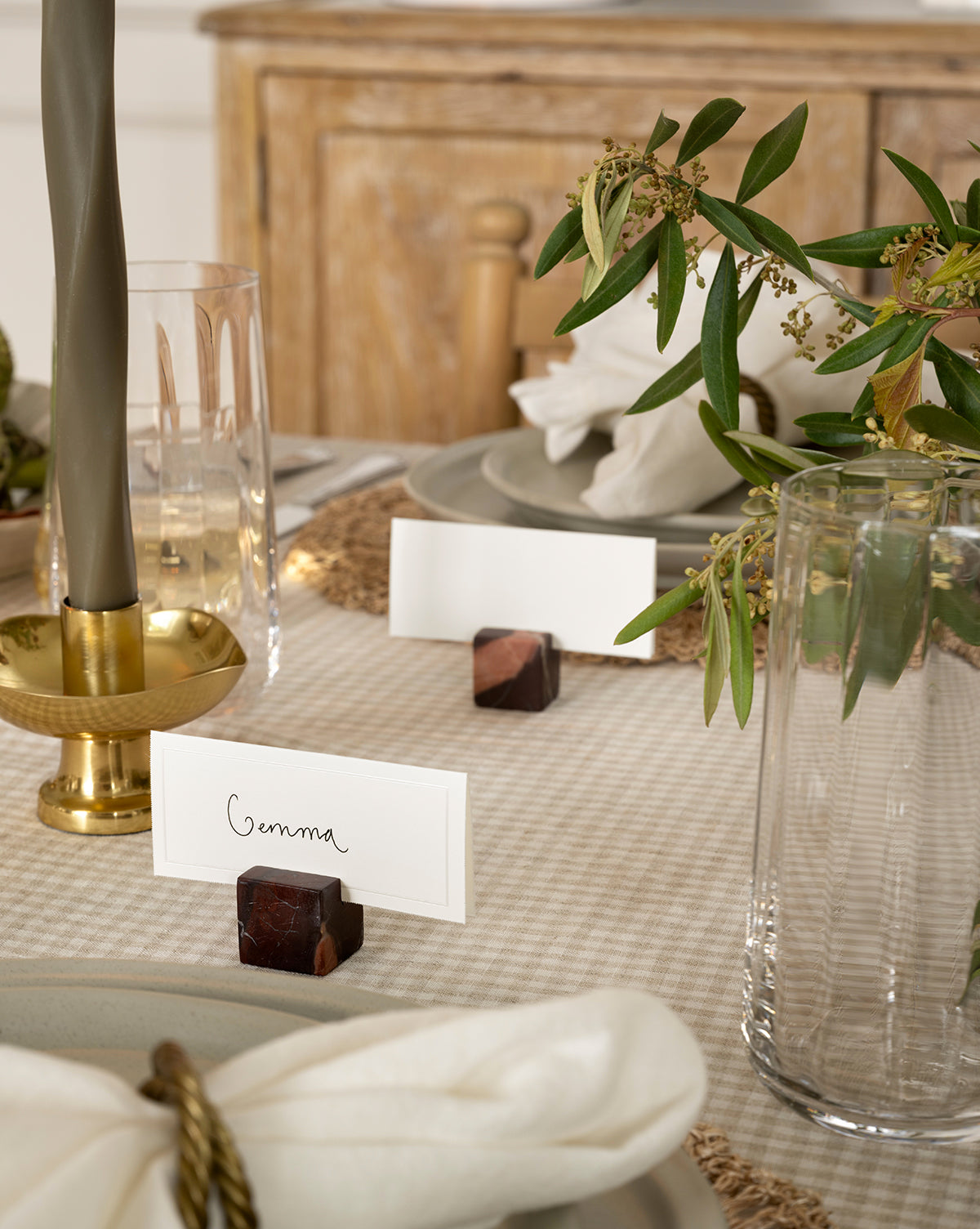 A close-up of an elegant harvest tablescape featuring the Auburn Marble Place Card Holders (Set of 4) with a name card, gold candlestick, clear glassware, white napkin, and green foliage on a beige checkered tablecloth—perfect for holiday gatherings, by Vendixs
