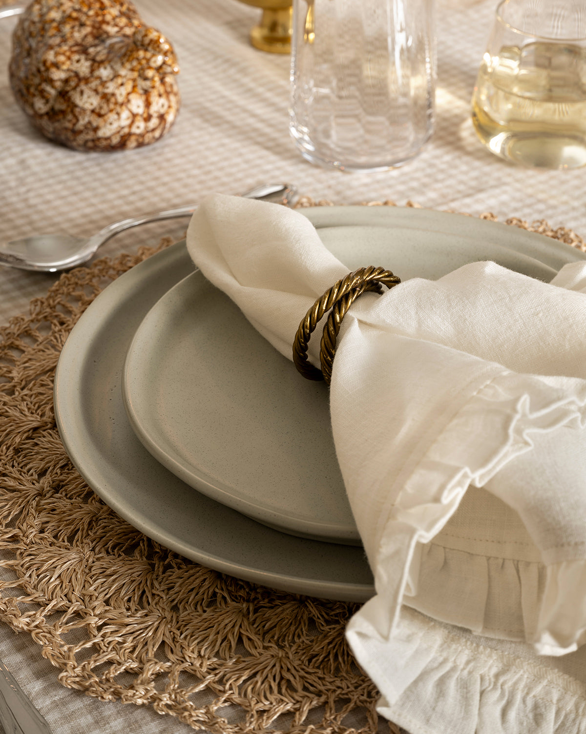 A close-up of a table setting featuring a cream-colored plate, the Aiden Soft Gray Side Plate, a linen napkin with a gold ring, and a woven placemat on a checkered tablecloth. A spoon, glasses, and decor are also visible by Vendixs