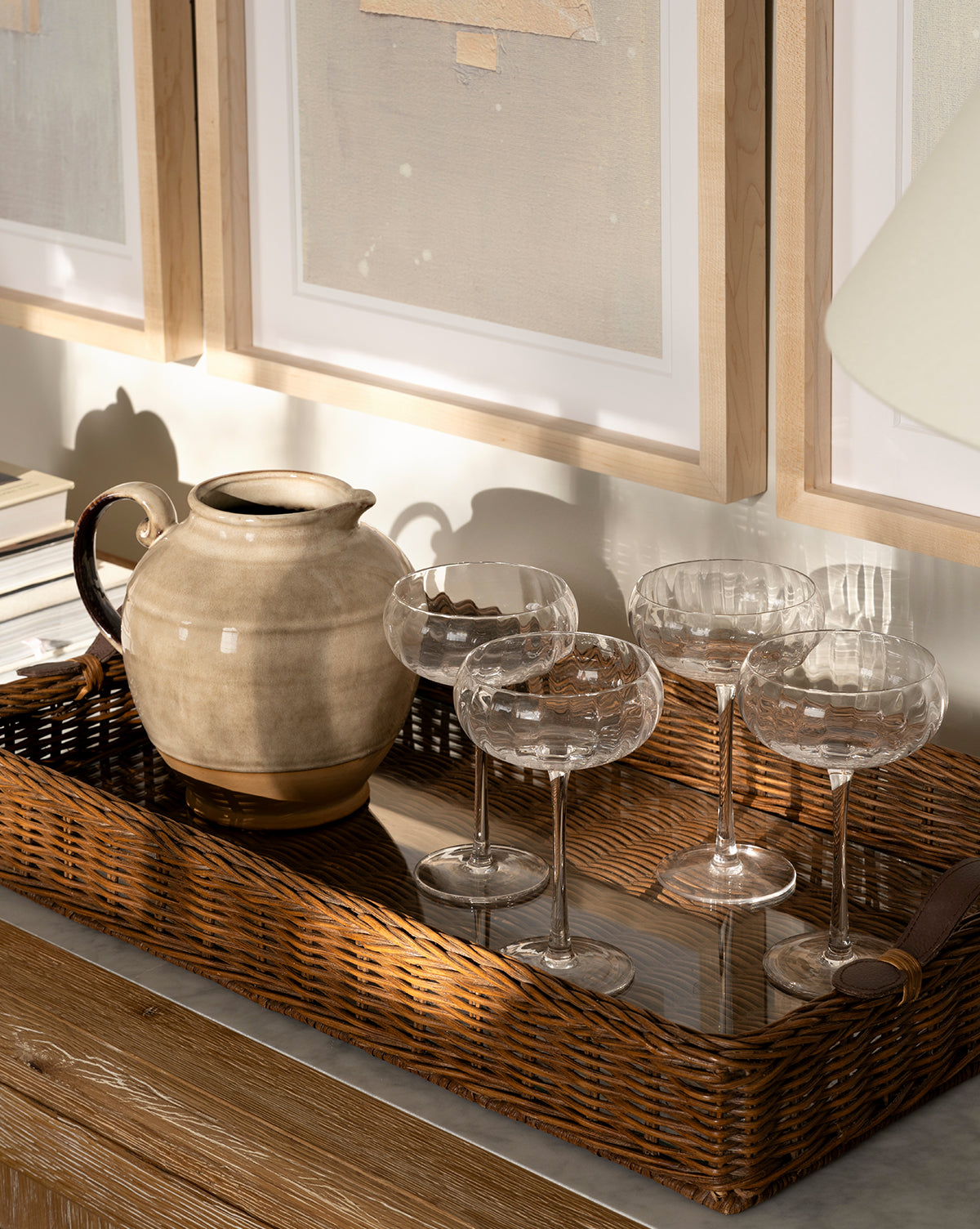 The Ashcroft Wicker Tray holds a beige ceramic pitcher and four coupe glasses on a wooden table, with framed abstract art displayed in the background, by Vendixs