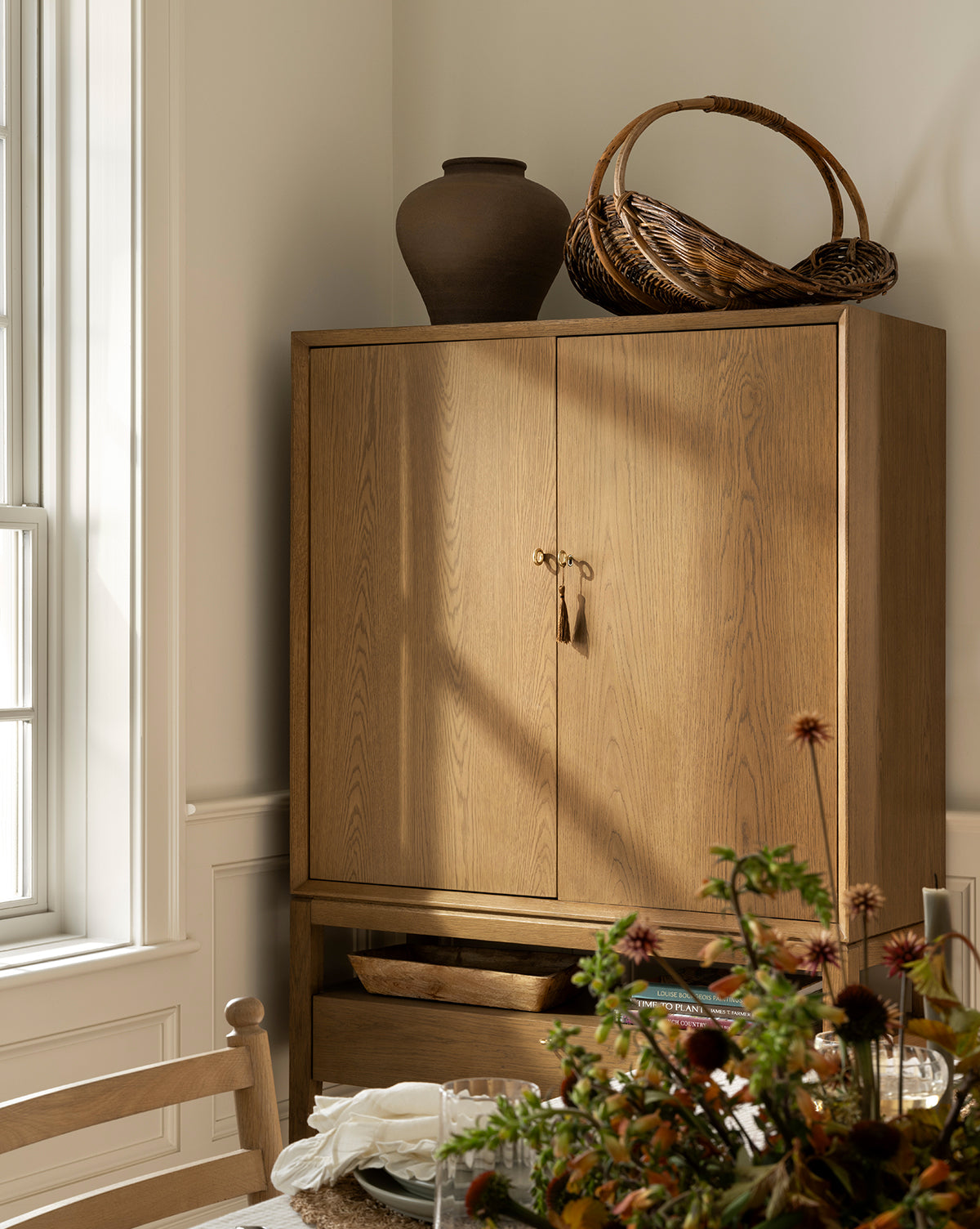 A wooden cabinet with closed doors stands in a sunlit room, topped with a matte brown Reading Vase and a woven basket. A dining table with flowers, napkins, and glassware is partially visible in the foreground, creating inviting home decor by Vendixs