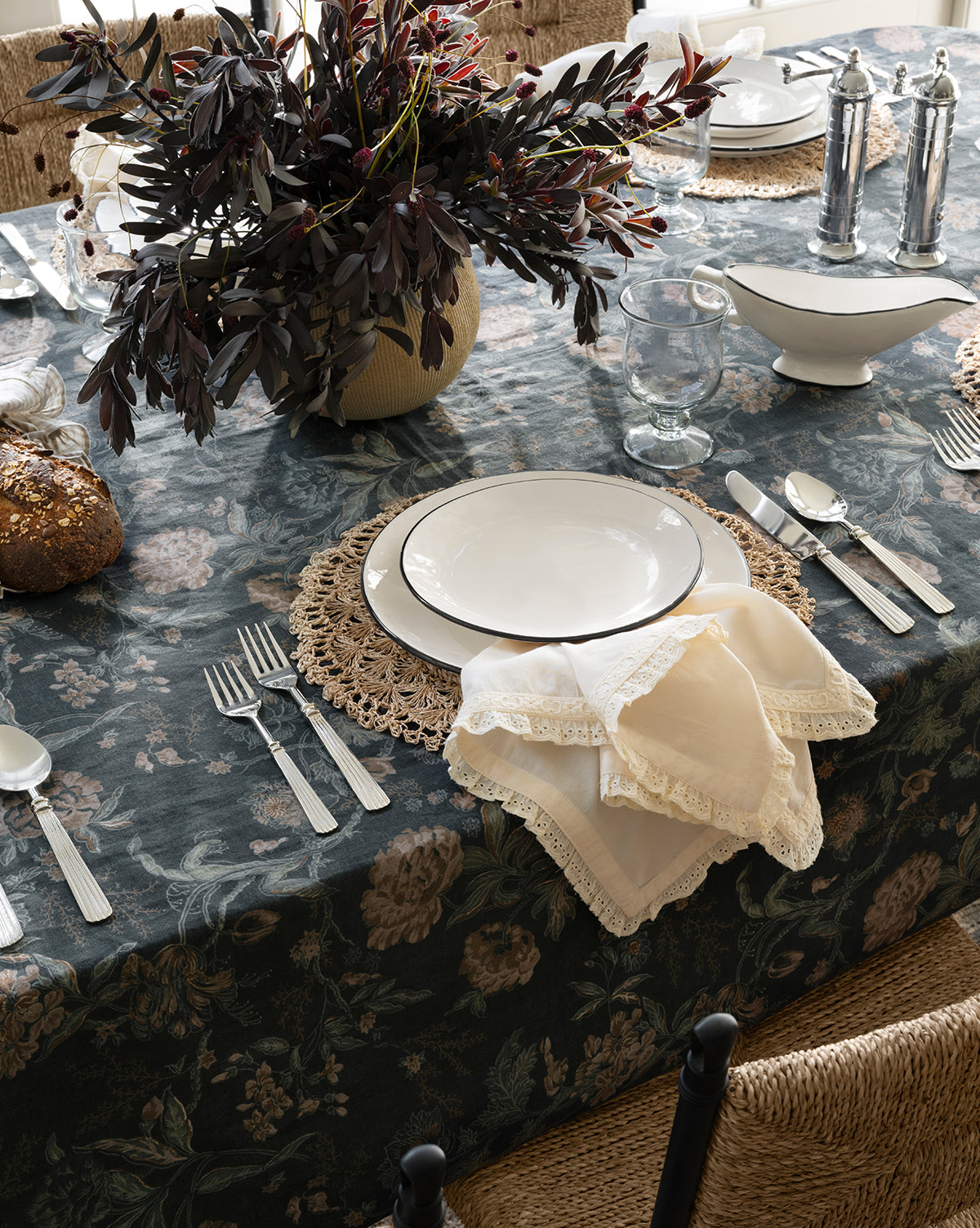 A dining table set with white plates, silver cutlery, lace-trimmed napkins, and glassware rests on the Hestercombe Tablecloth. A loaf of bread and a vase with leafy branches create a charming centerpiece, by Vendixs