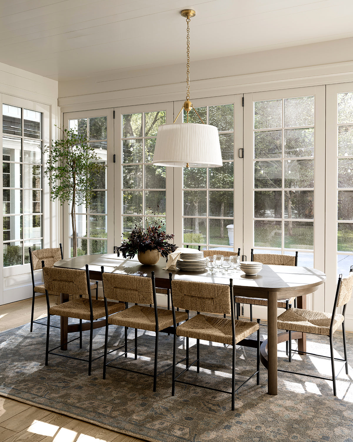 A bright dining room with a wooden table for eight, woven chairs, a large potted plant, and a white pendant light features tall windows and the Chevalier Hand-Knotted Wool Rug, adding timeless elegance to the space, by Vendixs