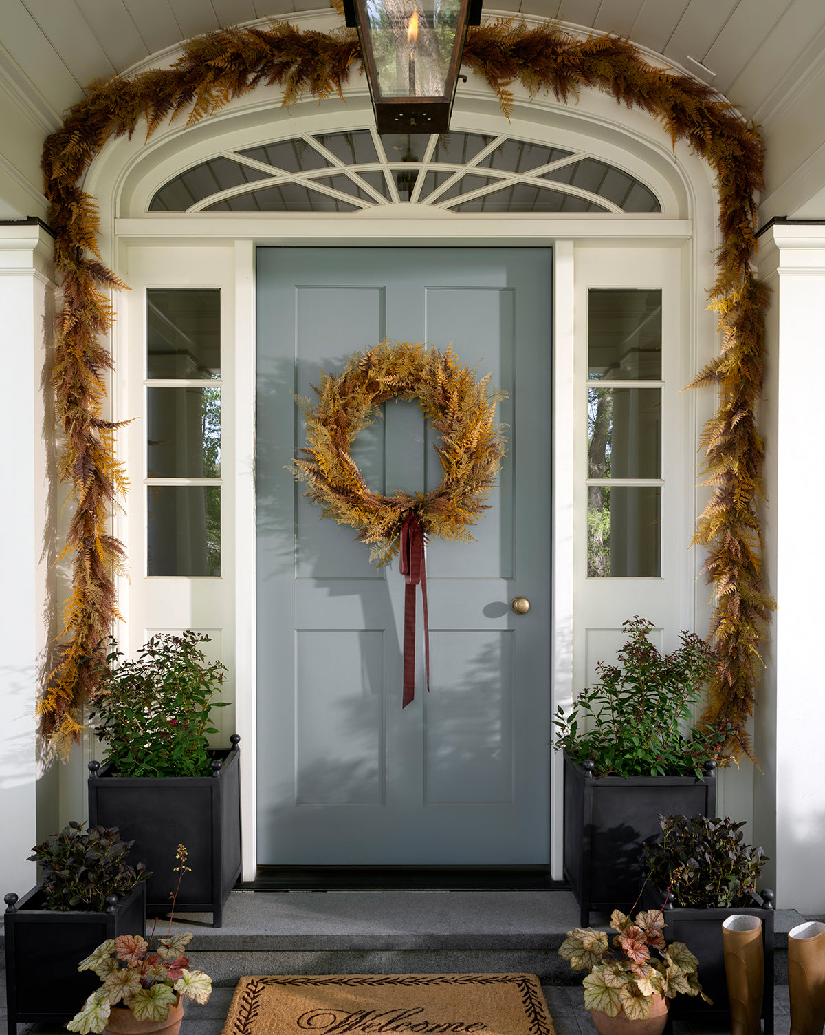 A blue front door displays the 30 Faux Golden Fern Wreath with coordinating garland, creating a stunning entryway. Black planters with leafy greens flank the Welcome doormat, all framed by elegant white columns, by Vendixs