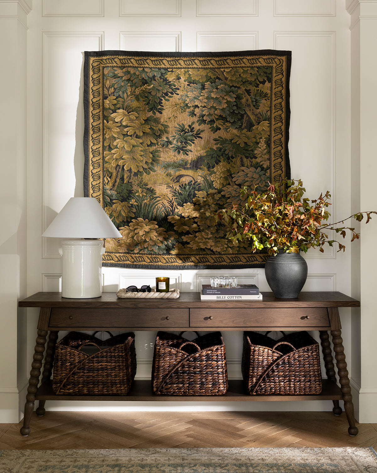 The Heathcliff Console, featuring a dark walnut oak finish and two drawers, sits against a white paneled wall topped by a green and gold tapestry. A white lamp, vase with branches, decor accents, and three woven baskets complete the look by Vendixs