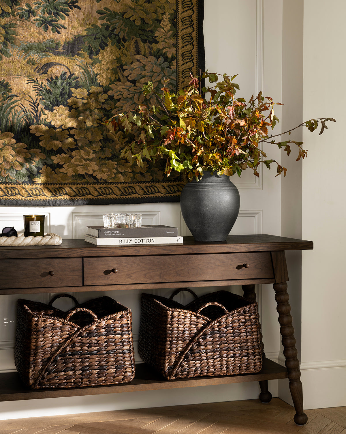 A dark wooden console table with a black vase, books, and candles sits below a tapestry. Two Quimby Baskets (Set of 2) are placed on the lower shelf against a white wall, by Vendixs