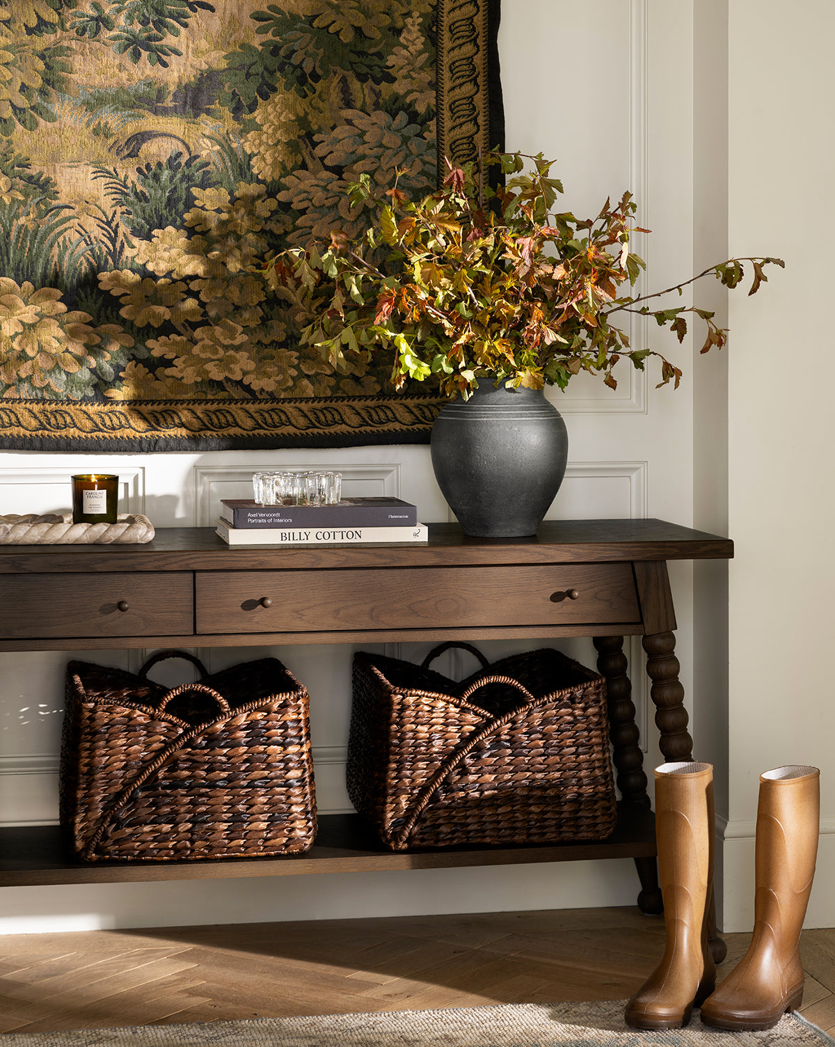 A wooden console table displays the Ethel Vase filled with autumn branches, alongside books and glassware. Woven baskets sit below, tall tan boots rest in front, and a tapestry provides rustic decor on the wall behind, by Vendixs