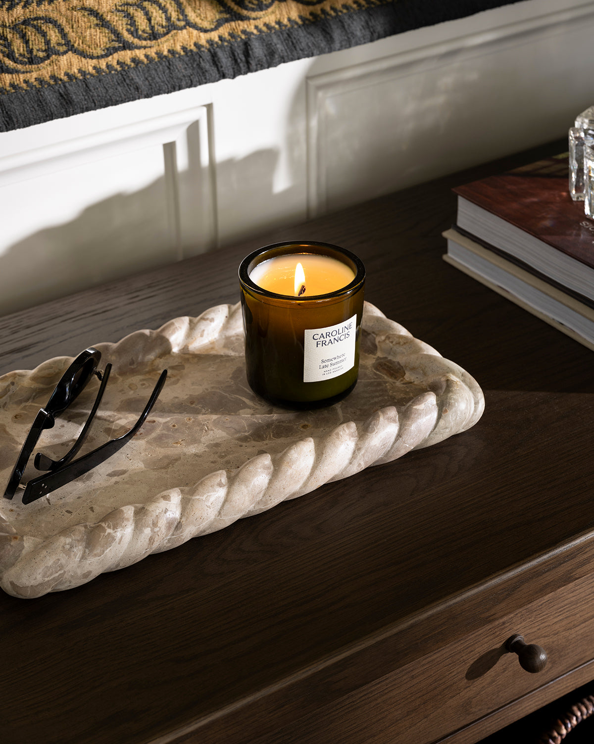 A lit scented candle in a glass jar sits on the Cleves Marble Tray next to eyeglasses on a dark wooden table, with books and a patterned bench adding cozy ambiance in the background, by Vendixs