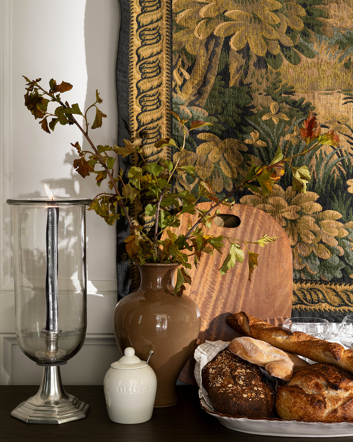 A decorative table features the Marlton Breadboard with assorted artisan breads, a tall candle in a glass holder, a brown vase with leafy branches, and a small ceramic jar, all set against a floral tapestry for artisanal kitchen decor by Vendixs