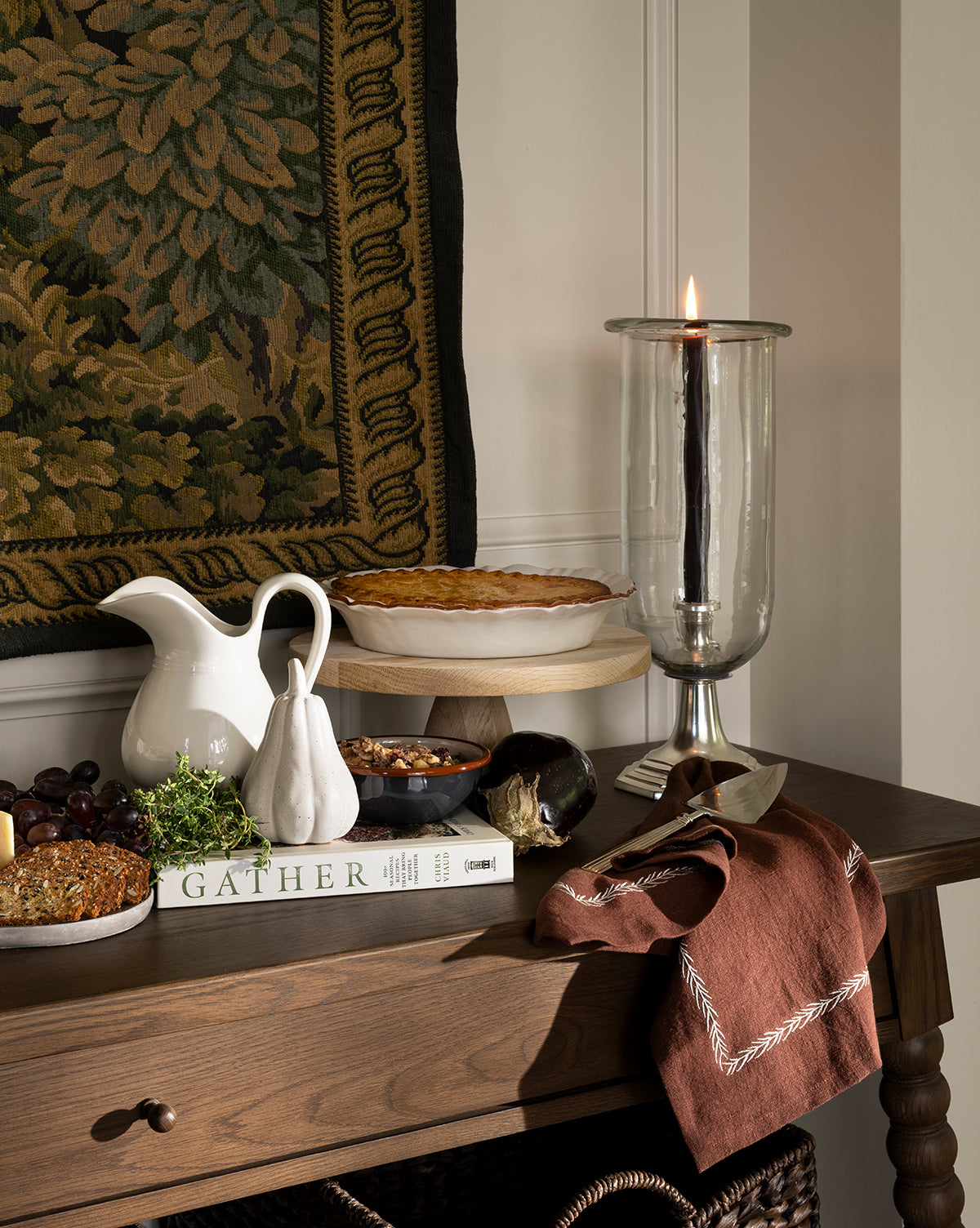 A wooden table features a pie, baked dish, eggplant, nuts, pear-shaped decor, brown napkin, Gather book, tall glass candleholder with lit black candle, and the white Lumen Pitcher set against a tapestry and white wall by Vendixs