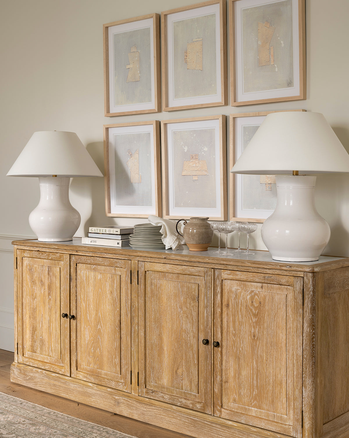 A wooden sideboard with four doors displays white lamps, books, dishes, glasses, and a ceramic jug. Above, Galactic IX by Jade Dacay adds striking minimalist style to the light-colored wall as modern wall decor by Vendixs