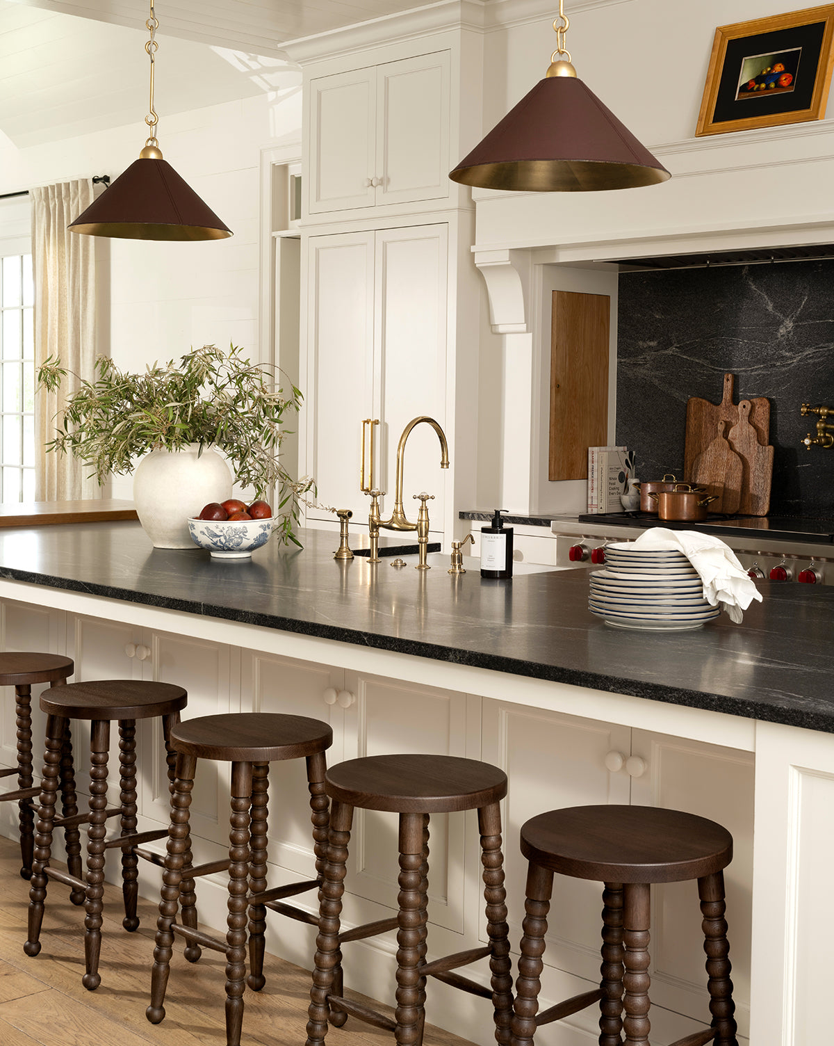A modern kitchen with a black countertop island, five dark wooden stools, gold fixtures, pendant lights, curated decor including stacked plates, a white vase with greenery, a bowl of apples, and Autumns Bounty I by Pascal Giroud art, by Vendixs