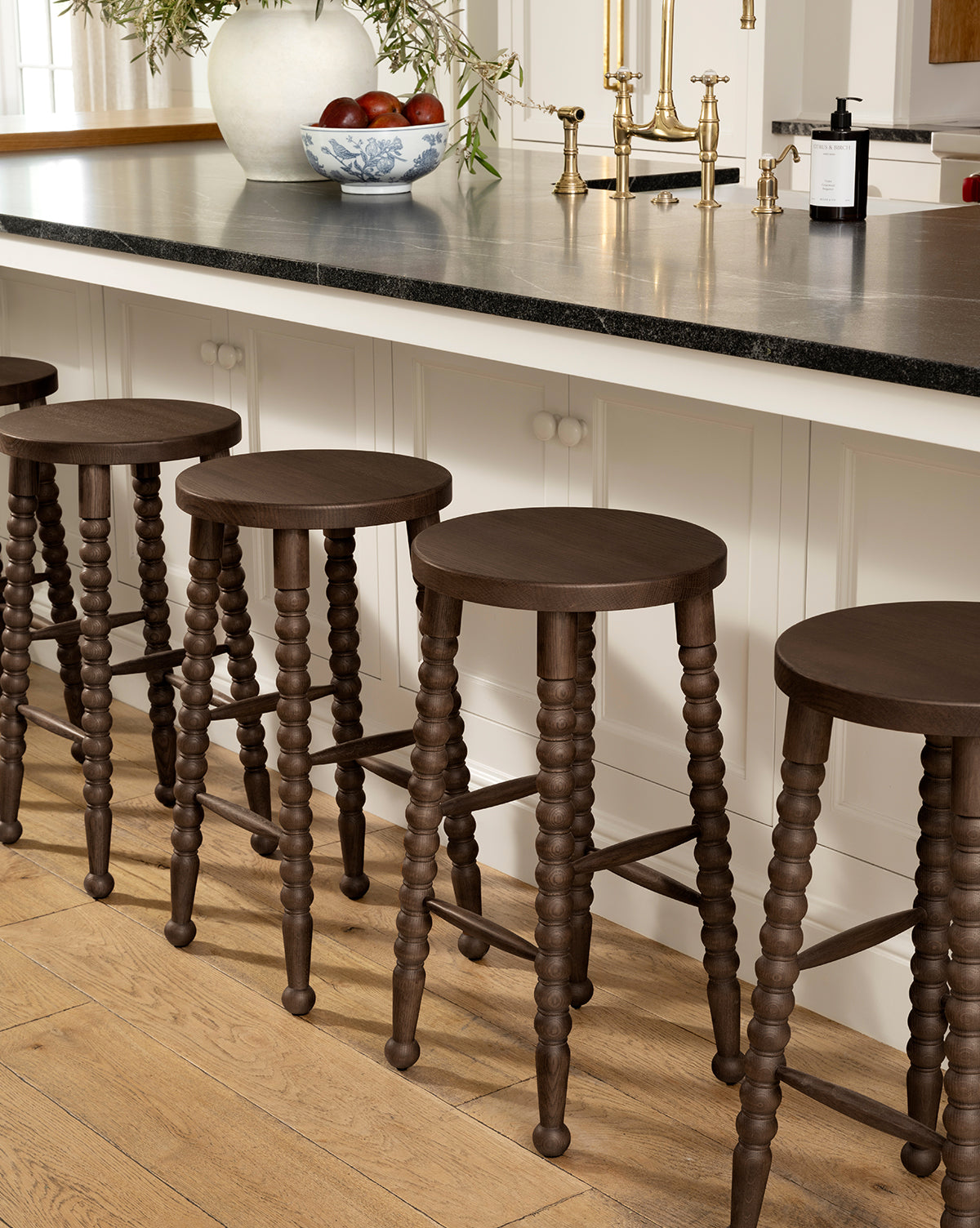 Five Heathcliff Stools with dark wooden, bobbin-style turned legs are lined up along a kitchen island topped with a black counter. A bowl of fruit, gold faucet, and soap dispenser sit on the counter; the floor is light wood, by Vendixs