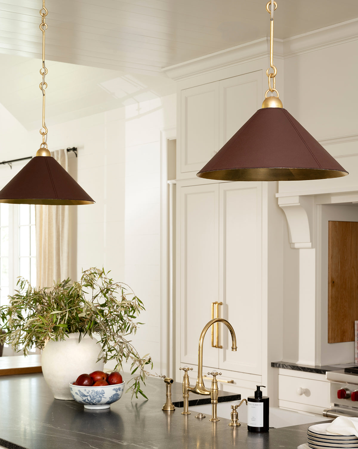 A bright kitchen featuring brass fixtures, a black countertop, elegant lighting from two Scarlett Pendants in brown and gold, a white vase with greenery, apples in a bowl, white cabinetry, and a large window by Vendixs