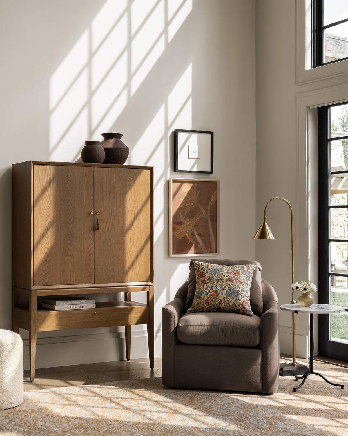 A cozy living room corner with a wooden cabinet, brown armchair and floral pillow, framed wall art, marble side table with flowers, sunlight streaming through tall windows, and the Vision Floor Lamp featuring a brass shade, by Vendixs