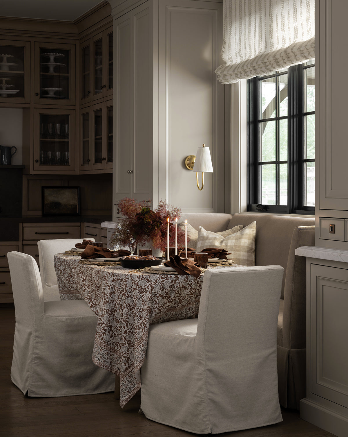 A cozy dining nook featuring four Olivier Slipcover Dining Chairs, a lace-covered table, a built-in bench with feather and down cushions, neutral cabinetry, candles, and a large window for a warm, inviting feel by McGee & Co.