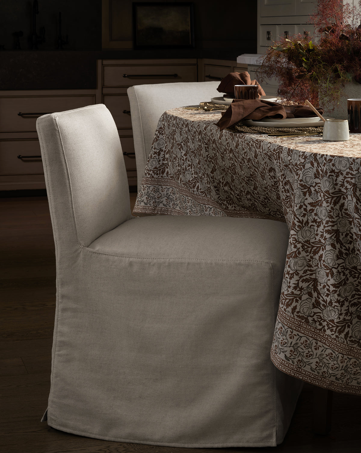 An Olivier Slipcover Dining Chair by Rowe Fine Furniture, Inc. sits next to a round table with a floral-patterned tablecloth, set with plates, cutlery, brown napkins, and a centerpiece of dried flowers, Vendixs