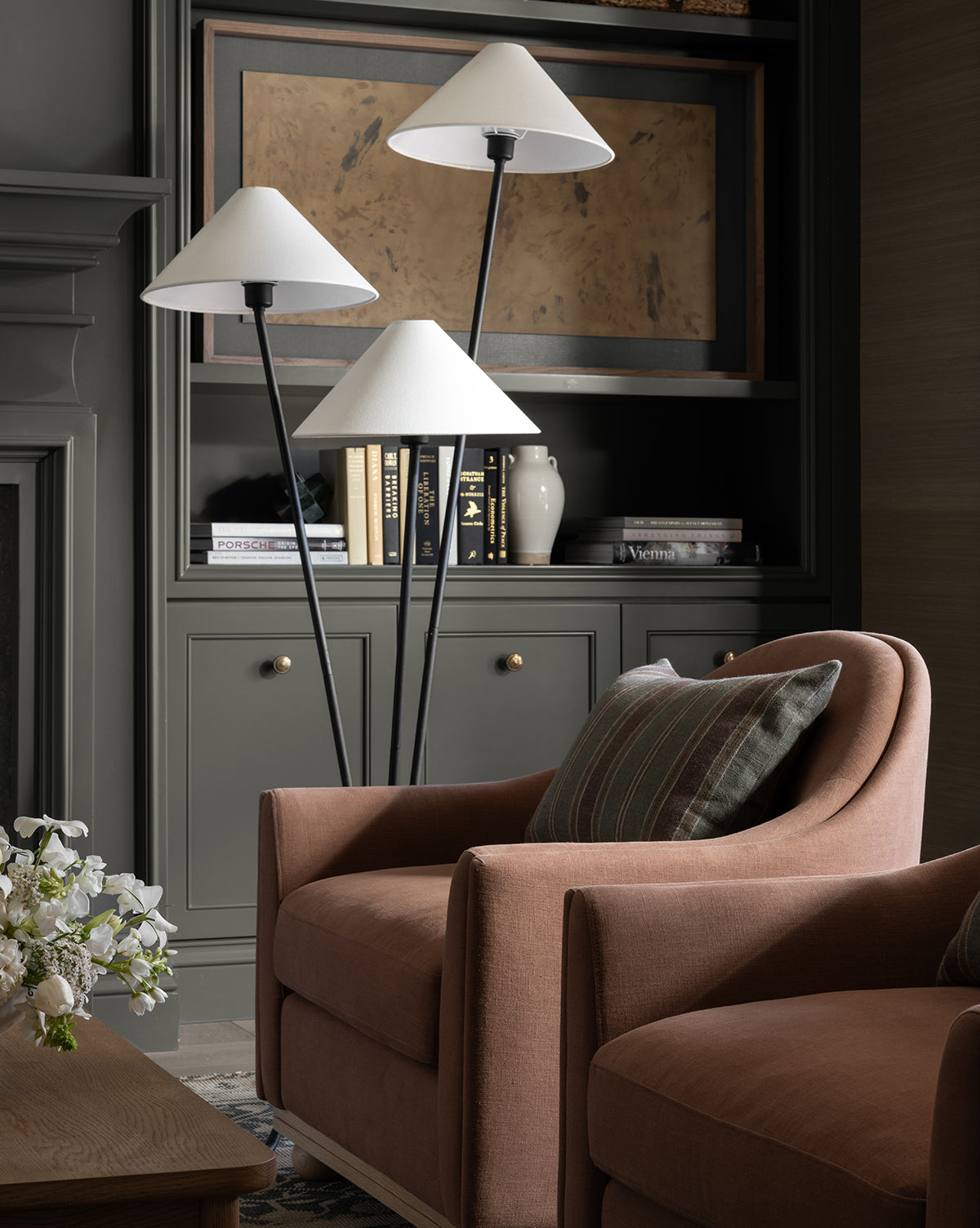A cozy living room with a soft brown armchair, striped pillow, and the Cedar Floor Lamp with linen shades. Built-in dark green shelves behind hold books, a vase, and framed artwork. White flowers rest on a wooden table, by Vendixs