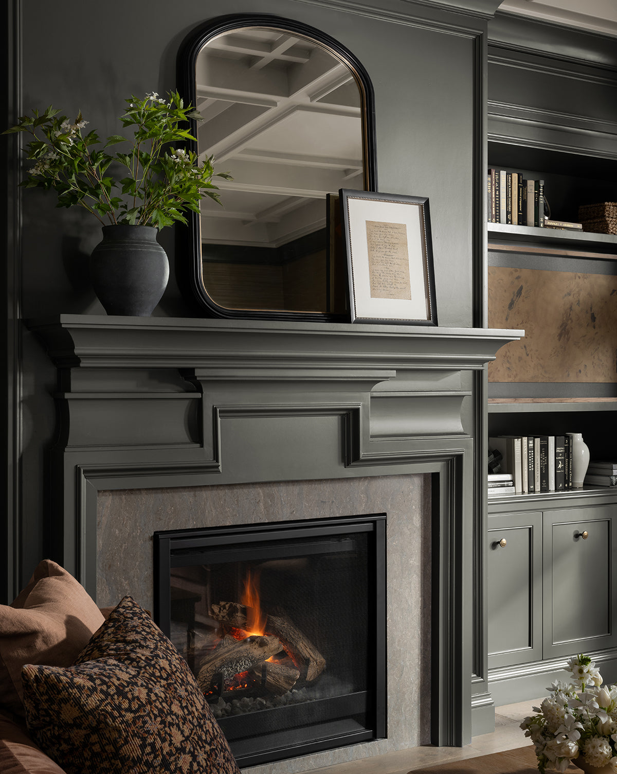 A stylish living room features a green-gray mantel with a fireplace, the Samuel Wall Mirror above it, a vase with green branches, a framed document, built-in shelves filled with books, and cozy pillows on the nearby sofa, by Vendixs
