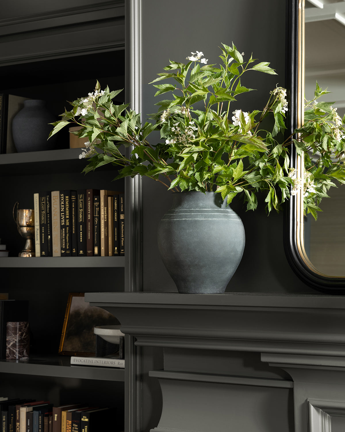 A dark gray room with built-in shelves, books, and the Ethel Vase—black terracotta filled with leafy branches and white flowers—on a mantel beneath a mirror, ideal for rustic home decor lovers, by Vendixs