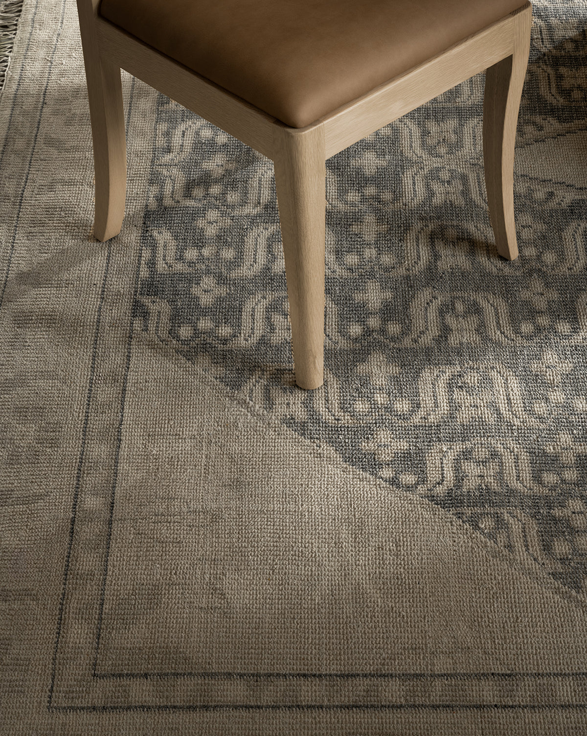 A close-up of a wooden chair with a tan seat cushion sits on the Elin Hand-Knotted Wool Rug, which displays geometric and floral motifs in muted gray and beige, showcasing its artisan craftsmanship, by Vendixs