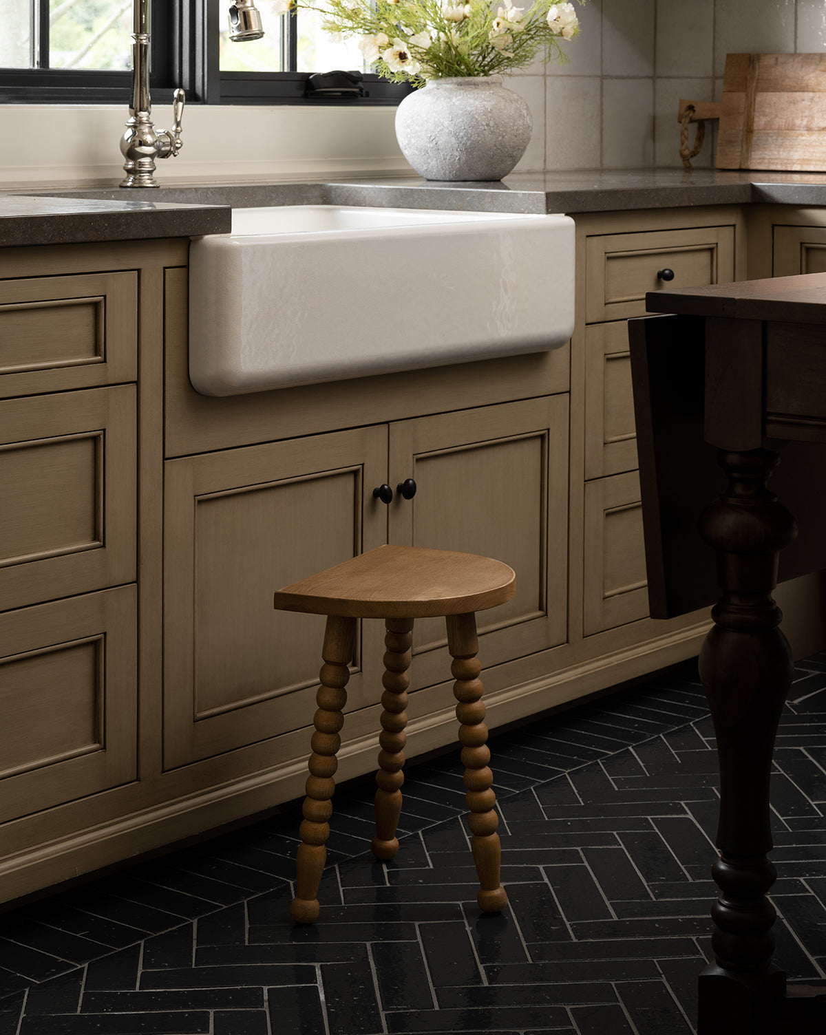 The Makerspalm Alice Honey Wood Stool with spindle legs sits before a farmhouse sink and beige cabinets in a kitchen featuring black herringbone tile flooring. A vase of flowers decorates the countertop by the window, Vendixs