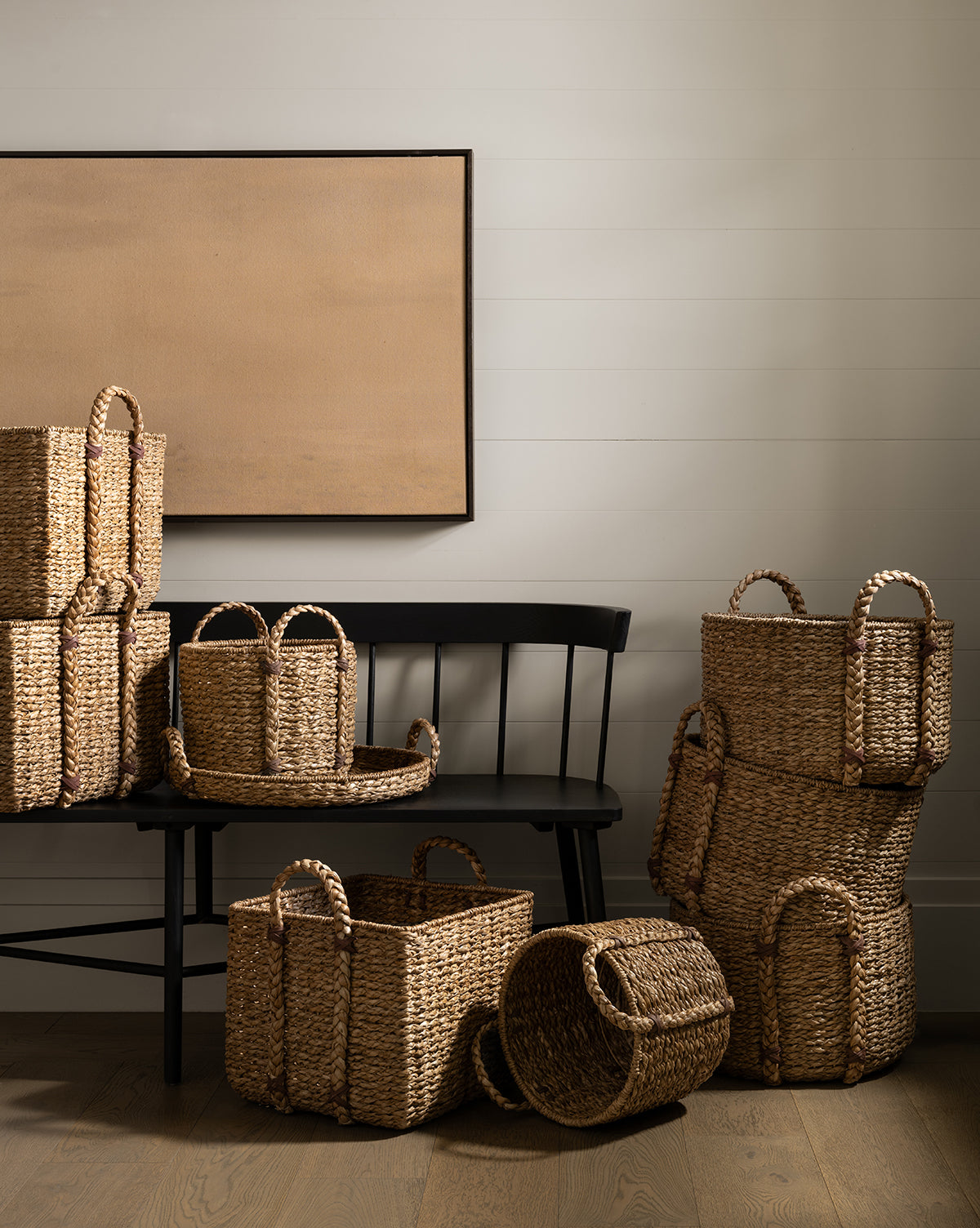 The Salter Woven Square Basket, along with other handled woven baskets, is displayed on and around a black wooden bench set against a white shiplap wall and beige canvas, adding an organized rustic touch to the wooden-floored space, by Vendixs